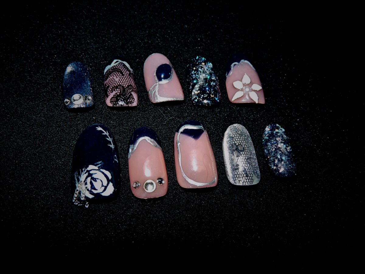 Untitled Ten short press-on nails in navy blue, nude pink, silver, and glitter finishes featuring lace, chrome outlines, rhinestones, floral art, textured glitter, and metallic details on a black background.
